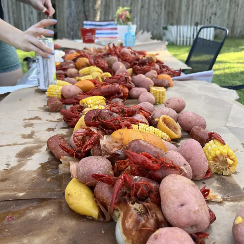 Long table covered in brown paper, piled with boiled crawfish, red potatoes, corn on the cob, sausage, onions and citrus halves, in a backyard