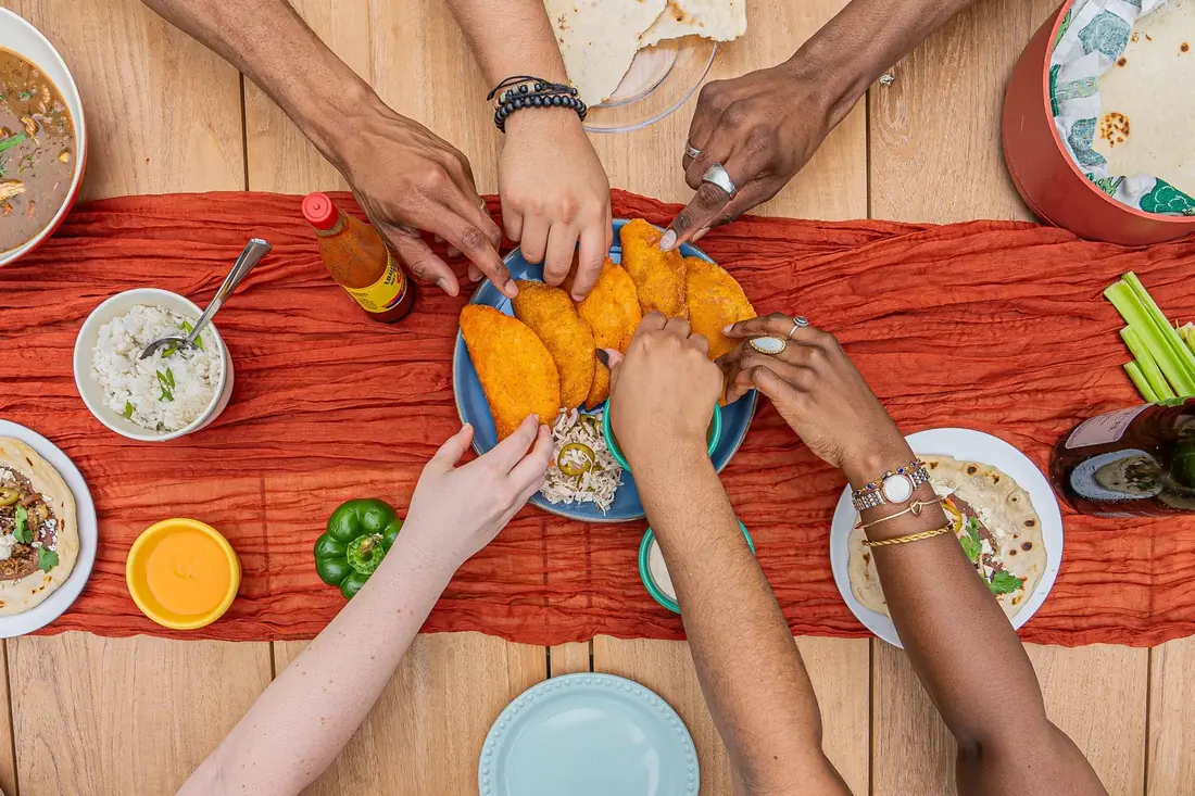Overhead view of a table with an orange runner: hands reach for golden fried turnovers on a blue plate, next to bowls of rice and soup, flour tortillas, a hot sauce bottle and a green pepper